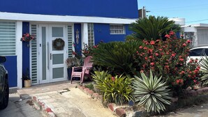 Una casa azul de dos pisos con una puerta y ventanas blancas, un camino de entrada y un pequeño jardín con flores rojas y una silla.