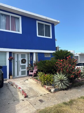 Ein zweigeschossiges blaues Haus mit weißer Tür und Fenstern, einer Auffahrt und einem kleinen Garten mit roten Blumen und einem Stuhl.
