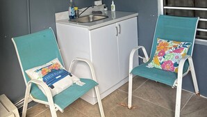 Una kitchenette pequeña con pileta y placares, dos sillas con cojines florales y un adorno de mariposa en la pared.