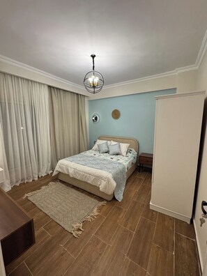 A bedroom with a bed, a nightstand, a dresser, a chandelier, and a window with curtains. - 3-Bedroom Apartment with AC, Near Grand Egyptian Museum, Wasal Compound. (Cairo Governorate)