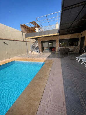 A swimming pool with a tiled deck, a covered outdoor seating area, and a staircase leading to a covered entrance.