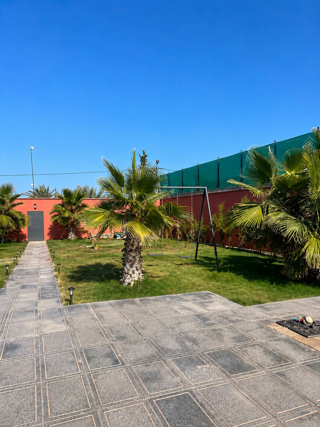 A paved pathway leading to a building with a red facade, surrounded by palm trees and a green fence.