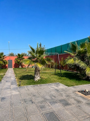 A paved pathway leading to a building with a red facade, surrounded by palm trees and a green fence.