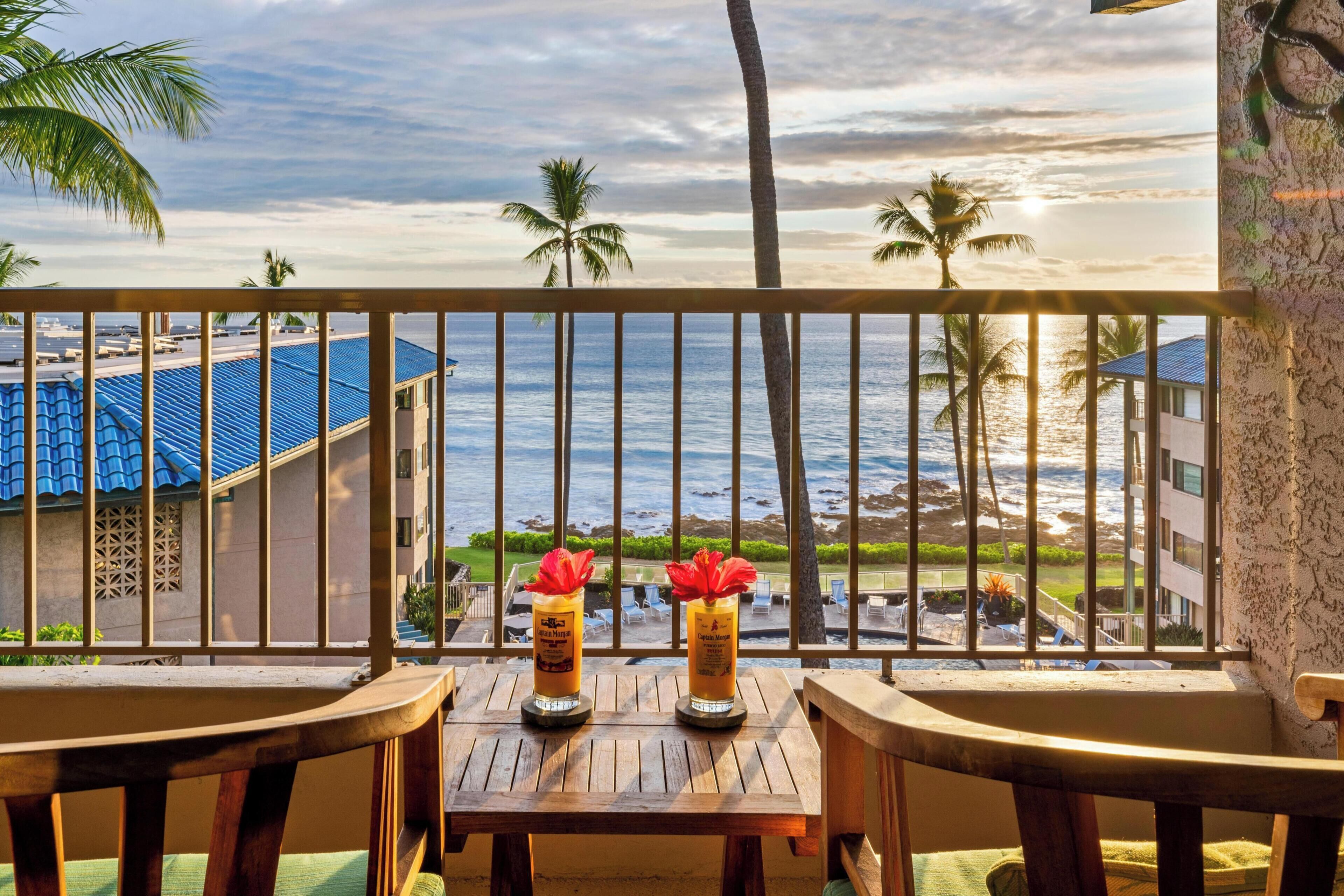 A balcony with two wooden chairs and a table, overlooking a coastal view with palm trees and a building with a blue roof.