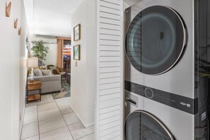 A laundry room with a front-loading washer and dryer, a wooden floor, and a hallway leading to a living area with a sofa, lamp, and framed pictures. - Kona Reef F22: Top-Floor Condo with Ocean Views (Kailua-Kona)