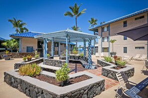 Tropical landscaping and pergola seating area at a beachside property, framed by palm trees and volcanic rock planters beneath a clear blue sky. - Kona Reef F22: Top-Floor Condo with Ocean Views (Kailua-Kona)