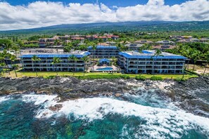 Oceanfront Kona Reef condo complex set on the Kona Coast shoreline with lush tropical grounds, swimming pool, and mountain backdrop. - Kona Reef F22: Top-Floor Condo with Ocean Views (Kailua-Kona)