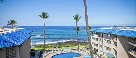 Lanai view showcasing the property's oceanfront location with a poolside courtyard, framed by tropical palms and blue tile roofs overlooking the Pacific shoreline.