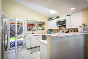 A kitchen with white cabinets, a microwave, a dishwasher, and a stone wall visible through an open door. - Hale Kona: Home - Ocean Views/Hot Tub/Private Yard (Kailua-Kona)