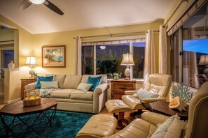 A living room with a beige sofa, a wooden side table, a blue rug, and a window with curtains. - Hale Kona: Home - Ocean Views/Hot Tub/Private Yard (Kailua-Kona)