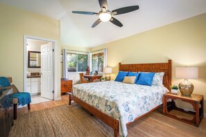 A bedroom with a bed, bedside tables, a nightstand, a ceiling fan, and a door leading to another room. - Hale Kona: Home - Ocean Views/Hot Tub/Private Yard (Kailua-Kona)