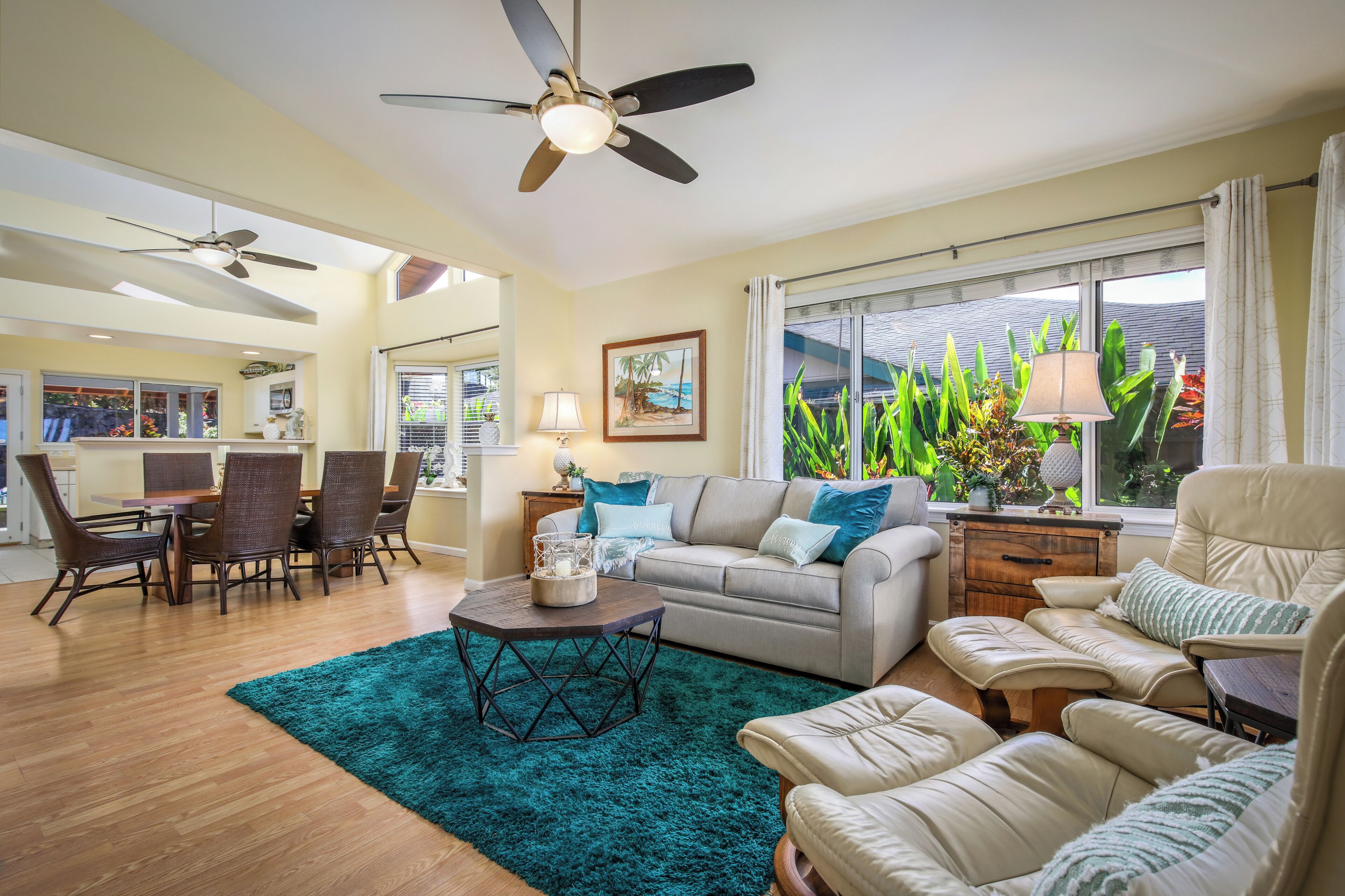 Un vaste salon doté d’un ventilateur de plafond, un coin repas avec des chaises en osier, un tapis turquoise et une vue sur des plantes tropicales par une grande fenêtre.