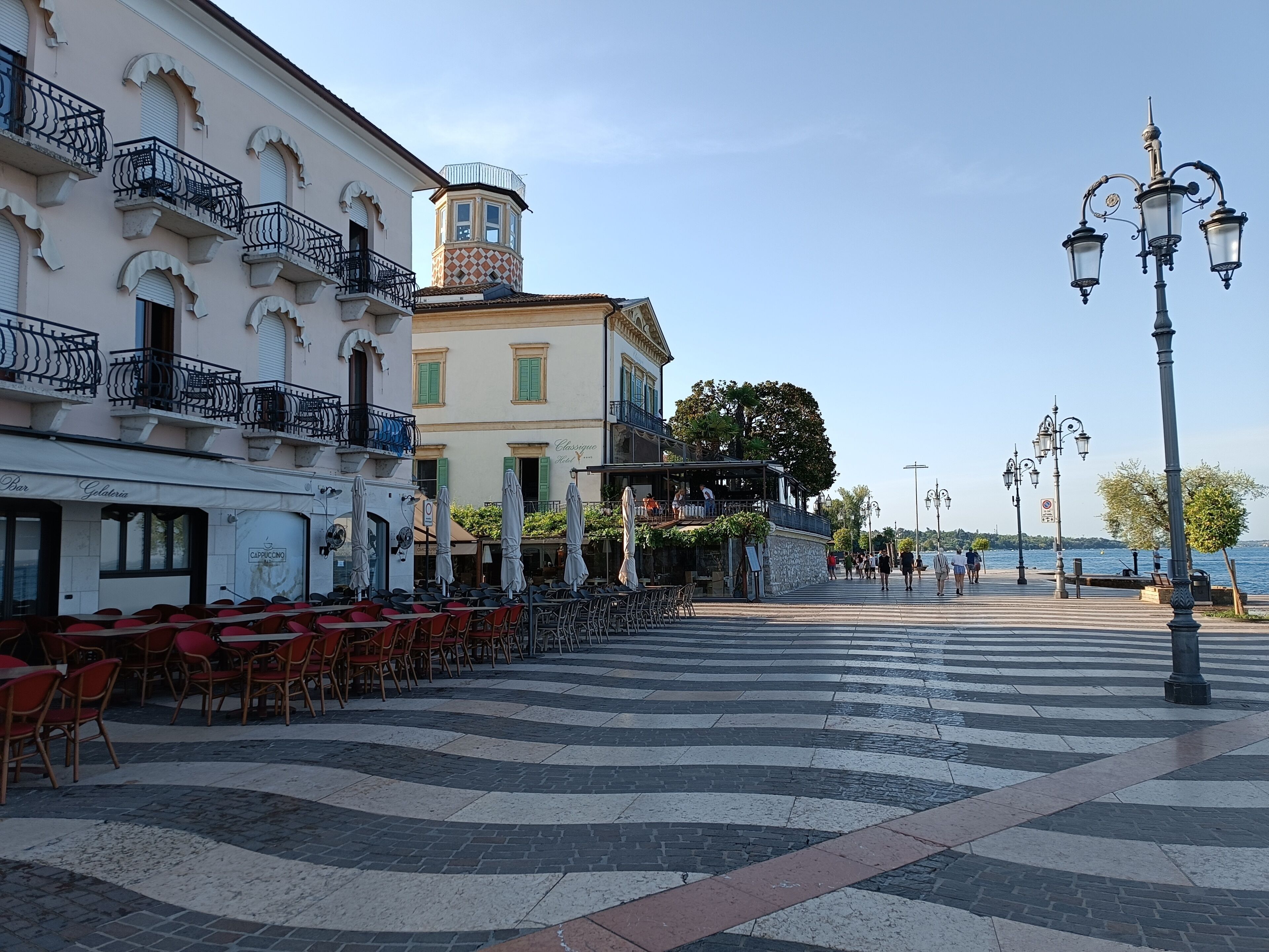 Un'area esterna con tavoli e sedie, vista sul lungomare e un edificio con balconi e una torre.