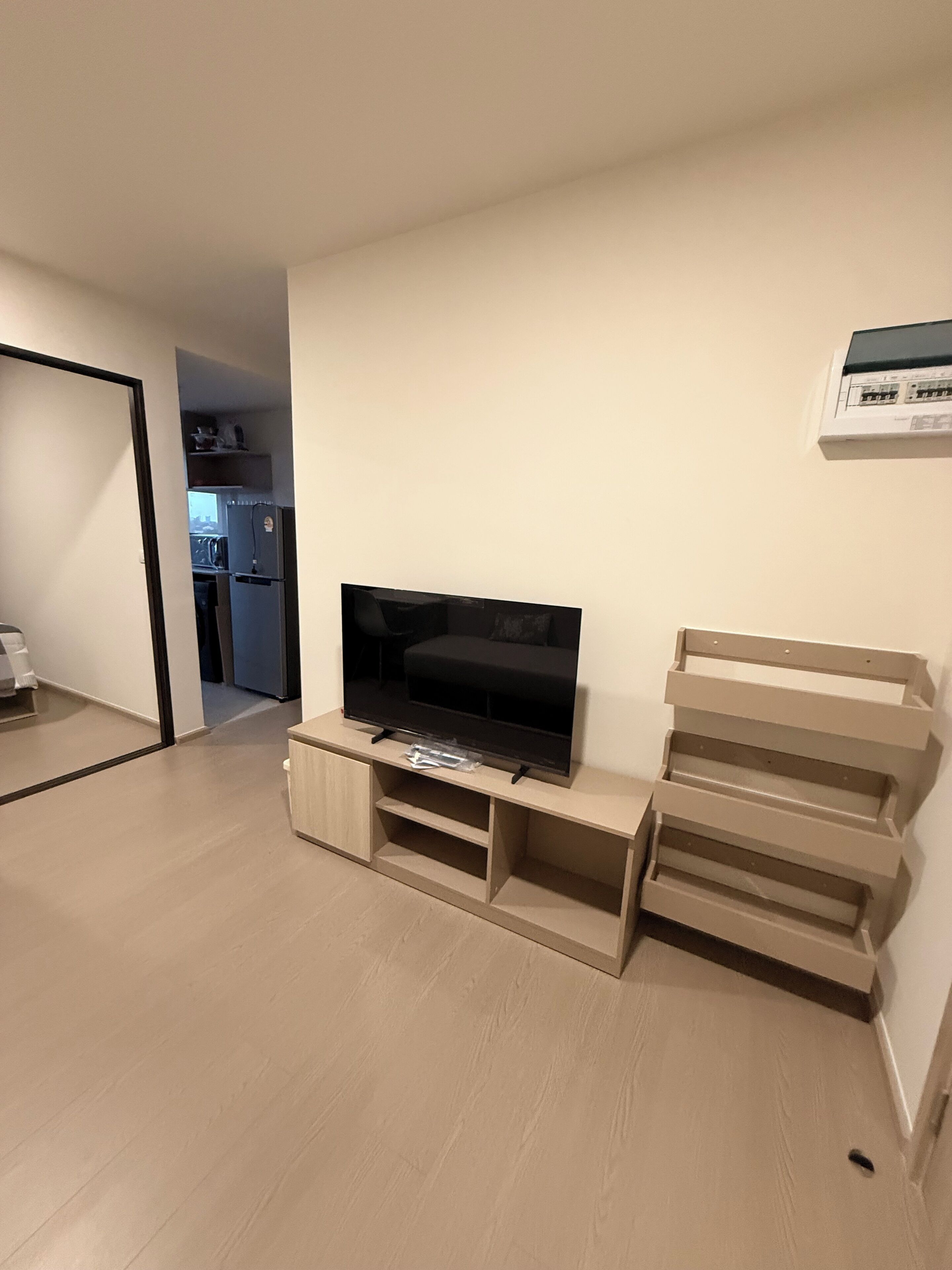 A living room with a flat-screen TV on a wooden stand, a kitchen visible through an open doorway, and a white wall-mounted air conditioning unit.