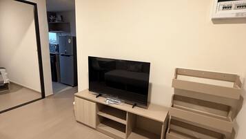 A living room with a flat-screen TV on a wooden stand, a kitchen visible through an open doorway, and a white wall-mounted air conditioning unit.