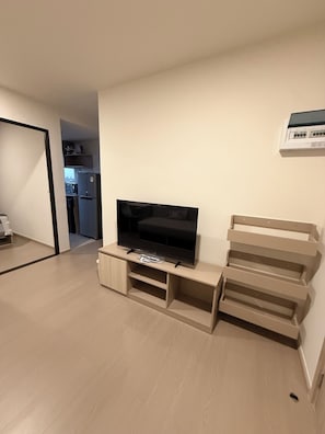 A living room with a flat-screen TV on a wooden stand, a kitchen visible through an open doorway, and a white wall-mounted air conditioning unit. - Cozy 1-bedroom apartment in fabulous Bang Chak area with AC (Krung Thep Maha Nakhon)