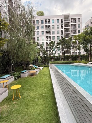 A modern outdoor pool area with a grassy lawn, seating, and a multi-story building in the background. - Cozy 1-bedroom apartment in fabulous Bang Chak area with AC (Krung Thep Maha Nakhon)
