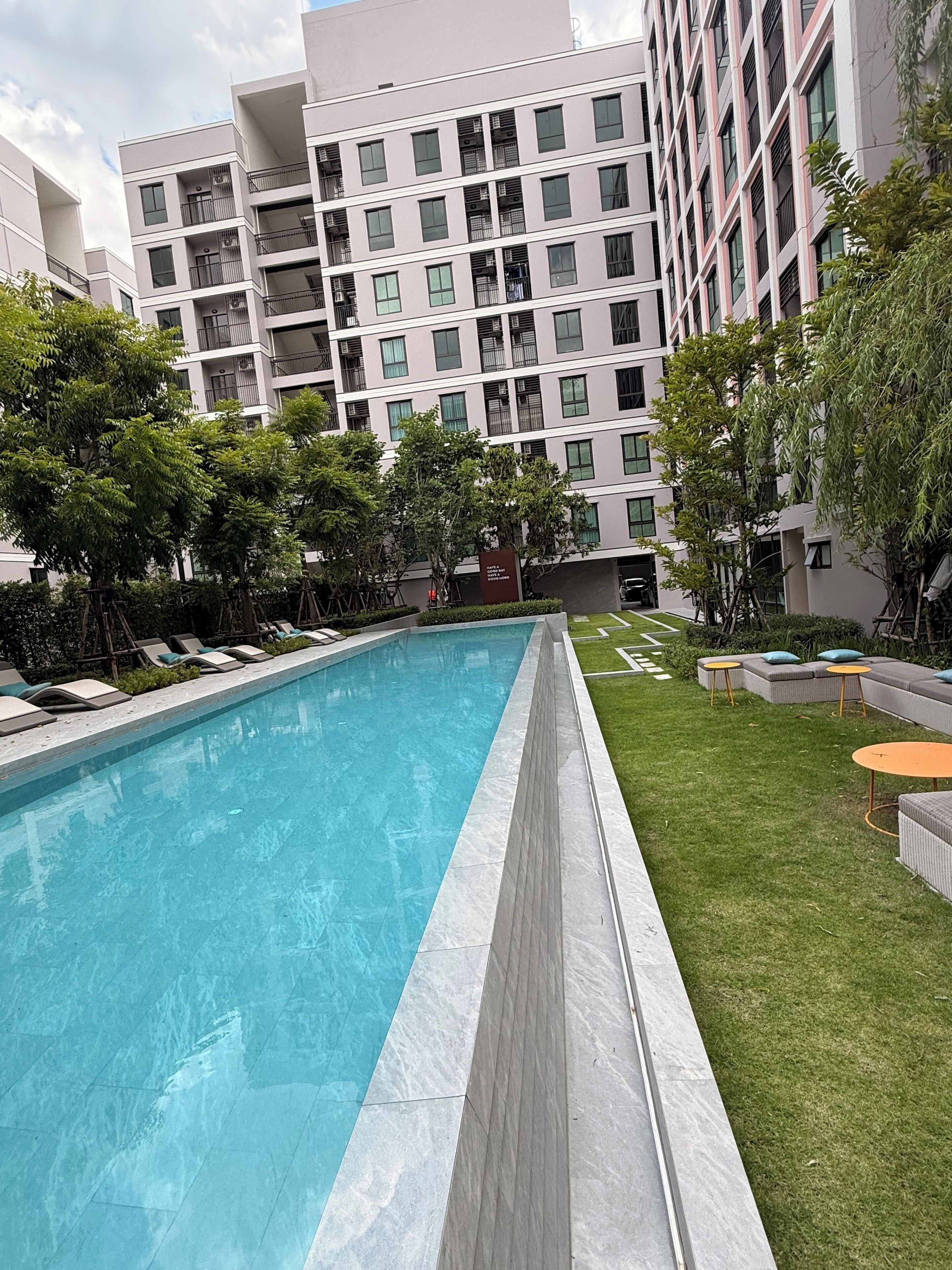 A swimming pool with a tiled edge, surrounded by grass and trees, with modern buildings in the background.