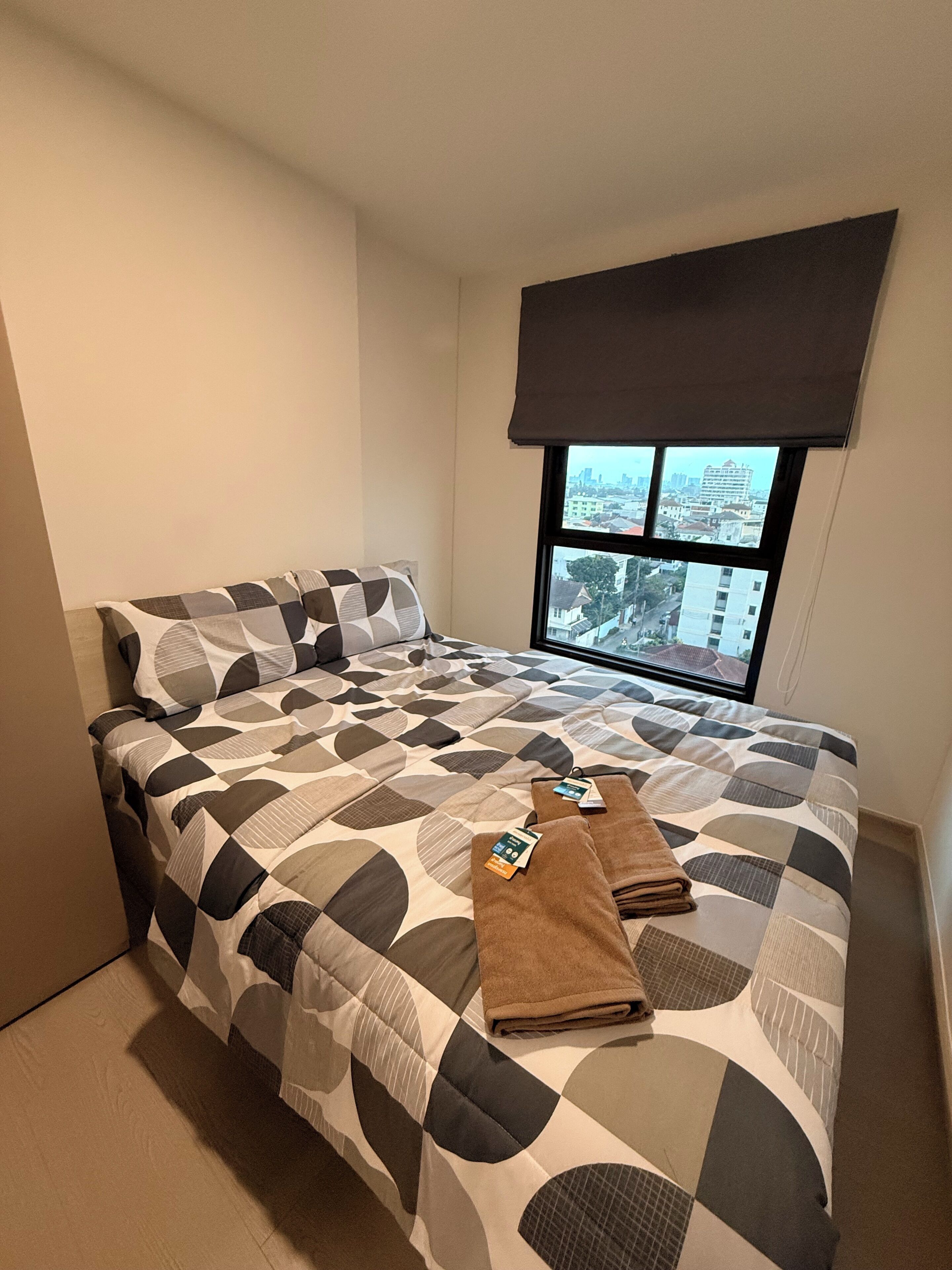 A single bed with a geometric patterned bedspread, a window with a city view, and a dark-colored headboard.