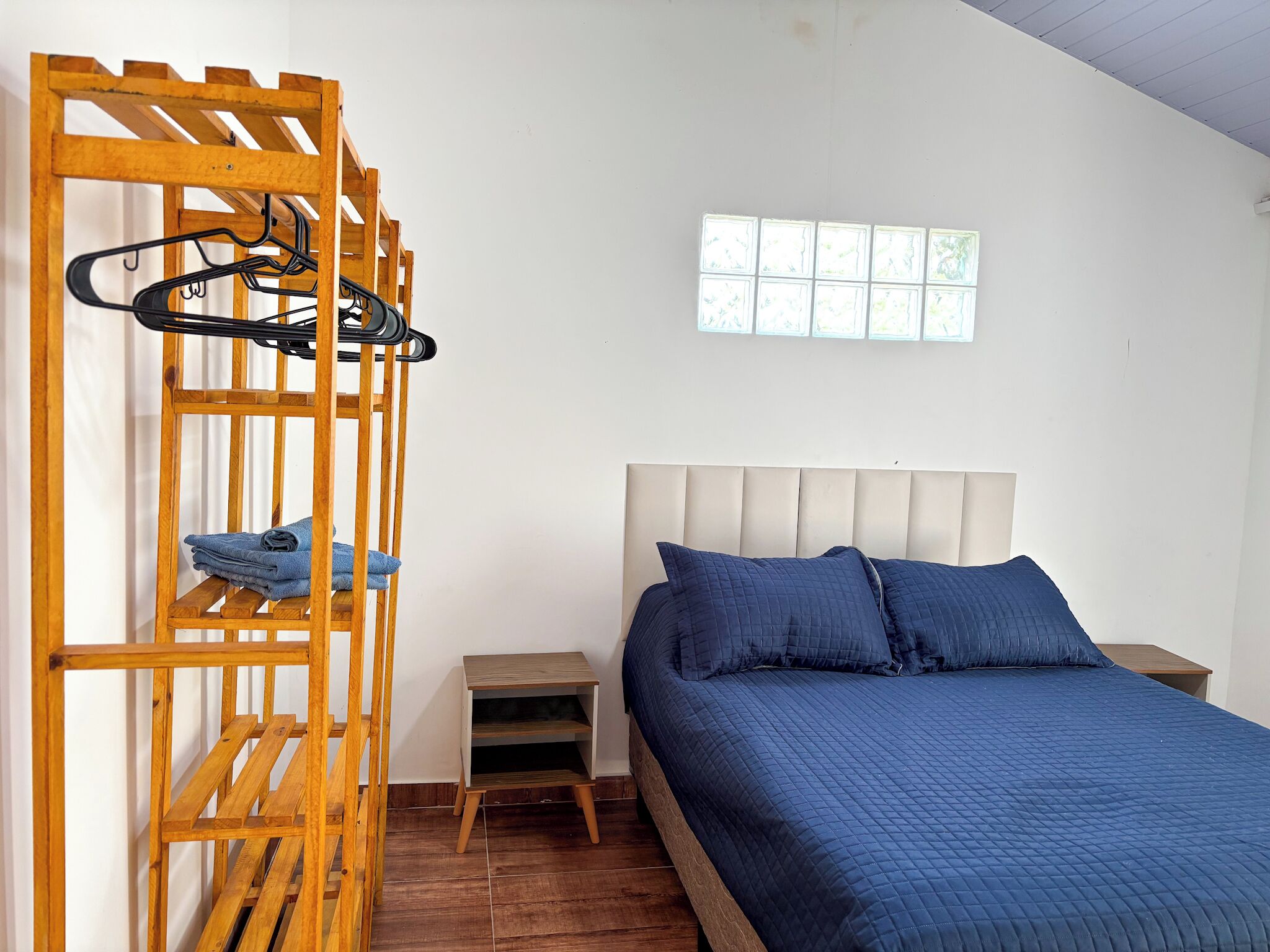 A bedroom with a bunk bed, a wooden clothes rack, a small bedside table, and a window with a view of greenery.