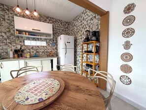 A kitchen with a white refrigerator, a wooden dining table with chairs, and a wall with geometric patterned tiles. - Casa Silandia Tartaruga, beaches, markets and city center within walking distance; excellent location. (Armação dos Búzios)