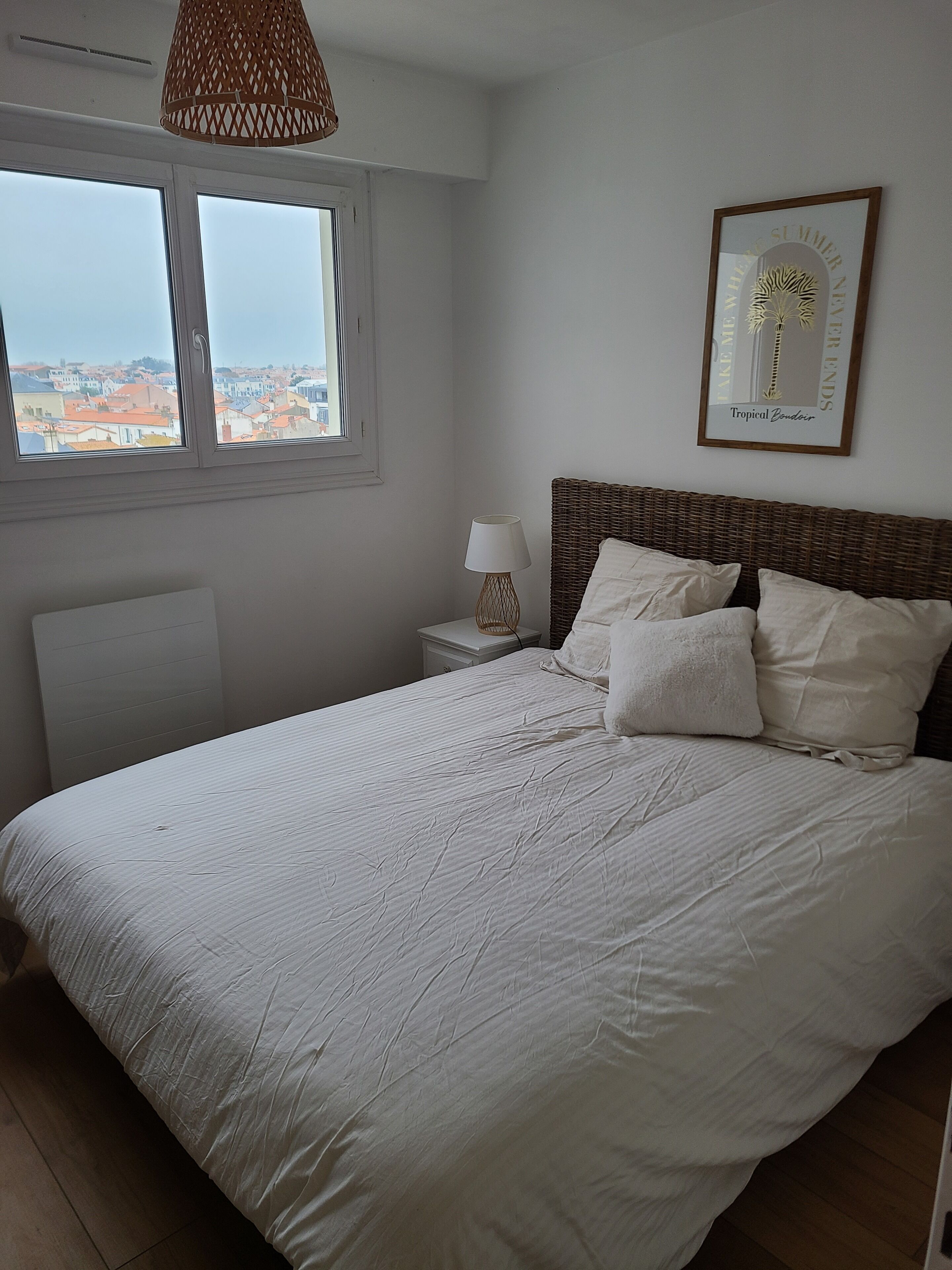 Une chambre avec un grand lit, une table de chevet avec une lampe, un tableau encadré au mur et une fenêtre donnant sur des immeubles.
