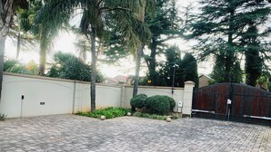 A paved driveway leading to a gated entrance with a white wall and a large tree.