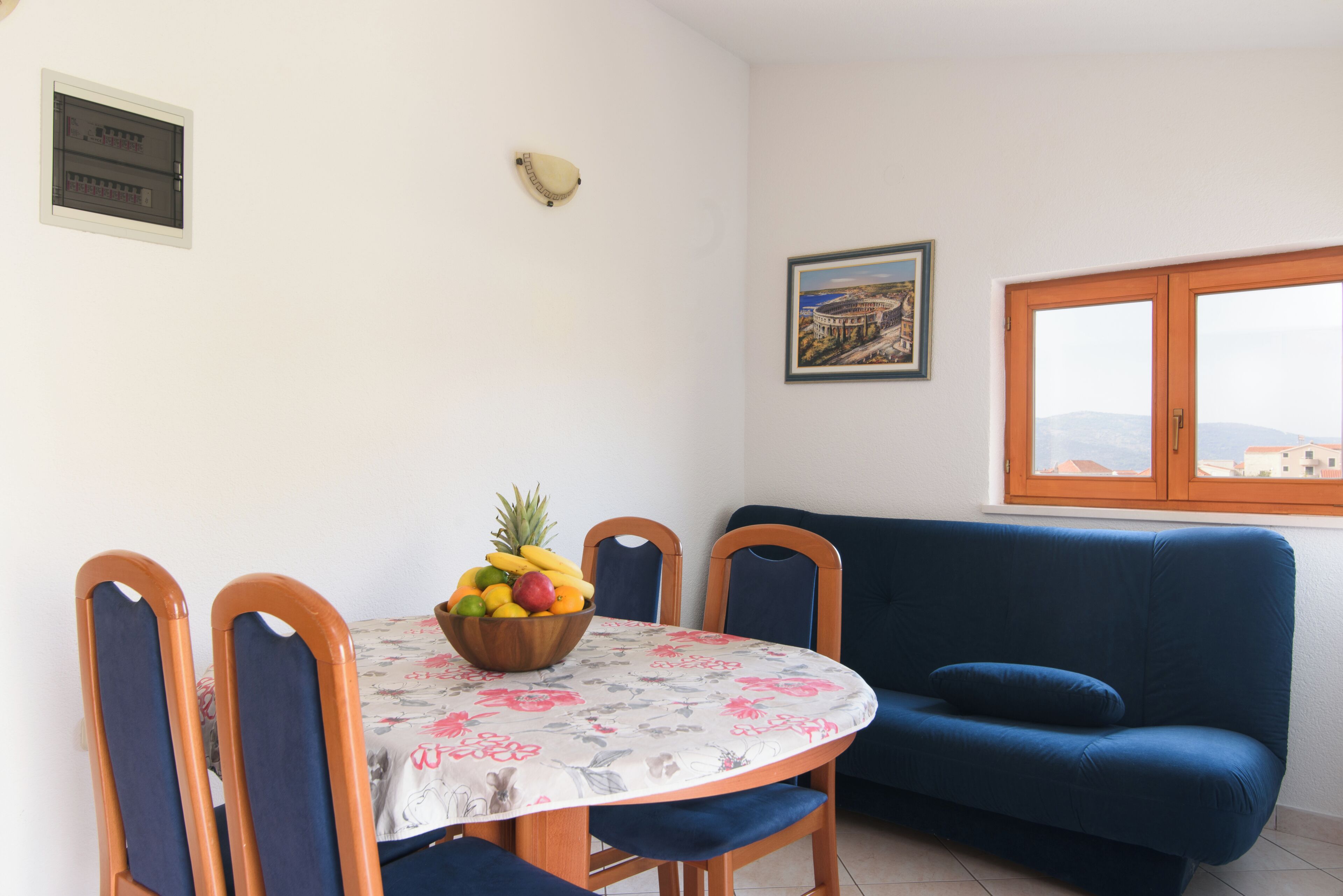 A dining area with a round table, chairs, a bowl of fruit, a couch, a window, and a framed picture on the wall.