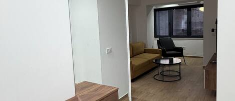 A modern living room with a sofa, a coffee table, and a chair. There is a wooden shelf and a large mirror on the wall.