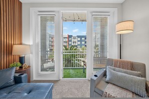 A modern living room with a sofa, a coffee table, and a lamp. There are two windows with blinds, and a view of buildings and greenery outside. - Modern 2BR w/ Pool + Pet-Friendly | Unit #24 (Tampa)