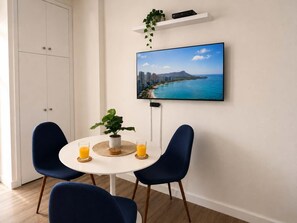 A dining area with a round table, two chairs, a potted plant, and a flat-screen TV mounted on the wall displaying a cityscape. - @ Marbella Lane - King Studio Steps to Beach Oasis (Honolulu)