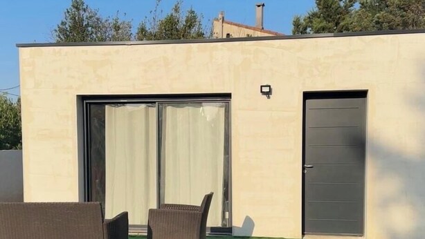 A modern building with a sliding glass door, a dark door, and two wicker chairs on a green artificial lawn.