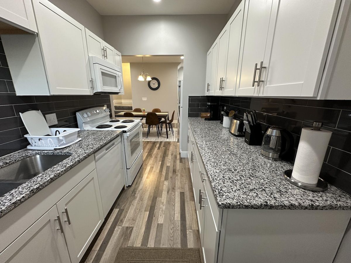 Una cocina moderna con armarios blancos, encimera de granito, estufa y un comedor con sillas y una mesa.