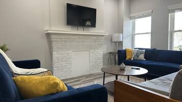 A living room with a fireplace, a blue sofa, a round coffee table, and a flat-screen TV mounted on the wall.