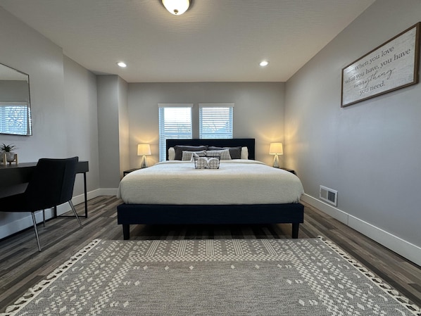 A bedroom with a bed, two bedside lamps, a desk with a chair, a rug, and a wall-mounted light fixture.