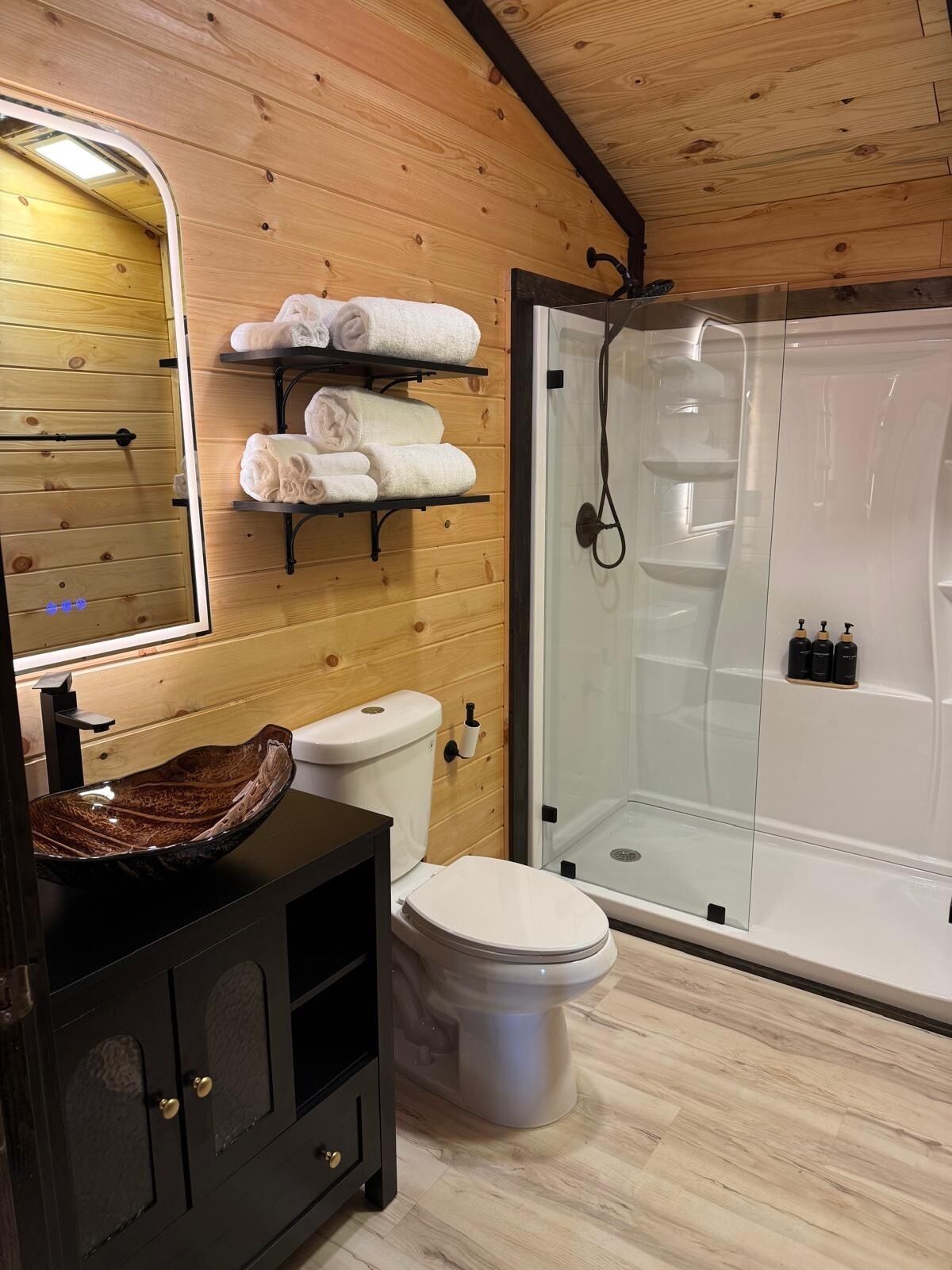 A bathroom with a wooden interior, a glass-enclosed shower, a sink with a bowl basin, and a toilet.