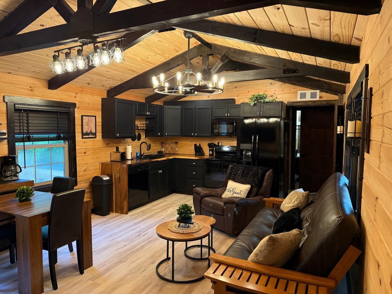 A cozy cabin interior with a dining area, a leather sofa, a small round table, and a kitchen with dark cabinets and a black refrigerator.