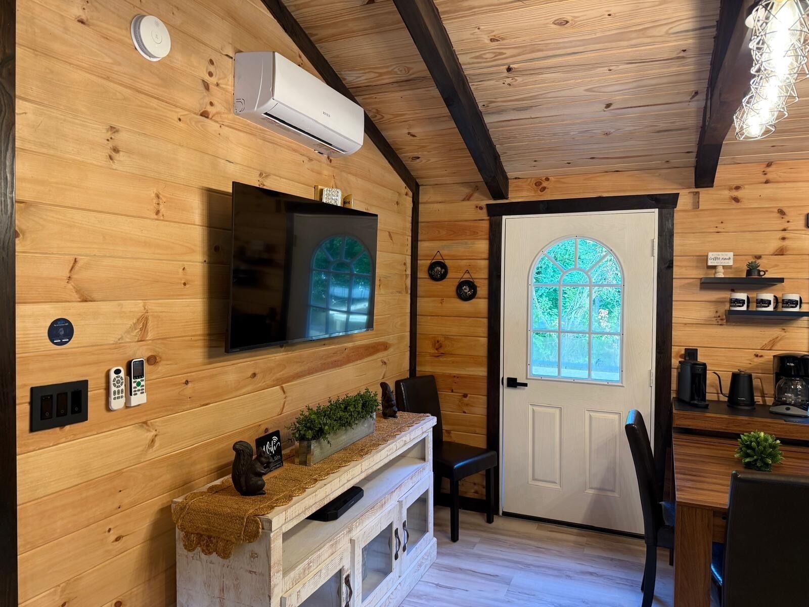 A wooden interior with a mounted air conditioner, a television, a door with a window, and a coffee station.