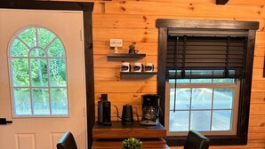 A wooden interior with a dining table, chairs, a coffee station, and a view through a window.