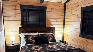 A wooden-paneled room with a bed, two bedside tables, and a window with shutters.