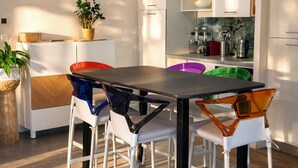 A modern kitchen with white cabinets, a wooden ceiling beam, and a dining area with colorful chairs. - Apartment in a renovated farmhouse: 2 bedrooms · 40 m² terrace, garden, parking · 12 km from Toulouse (Lapeyrouse-Fossat)