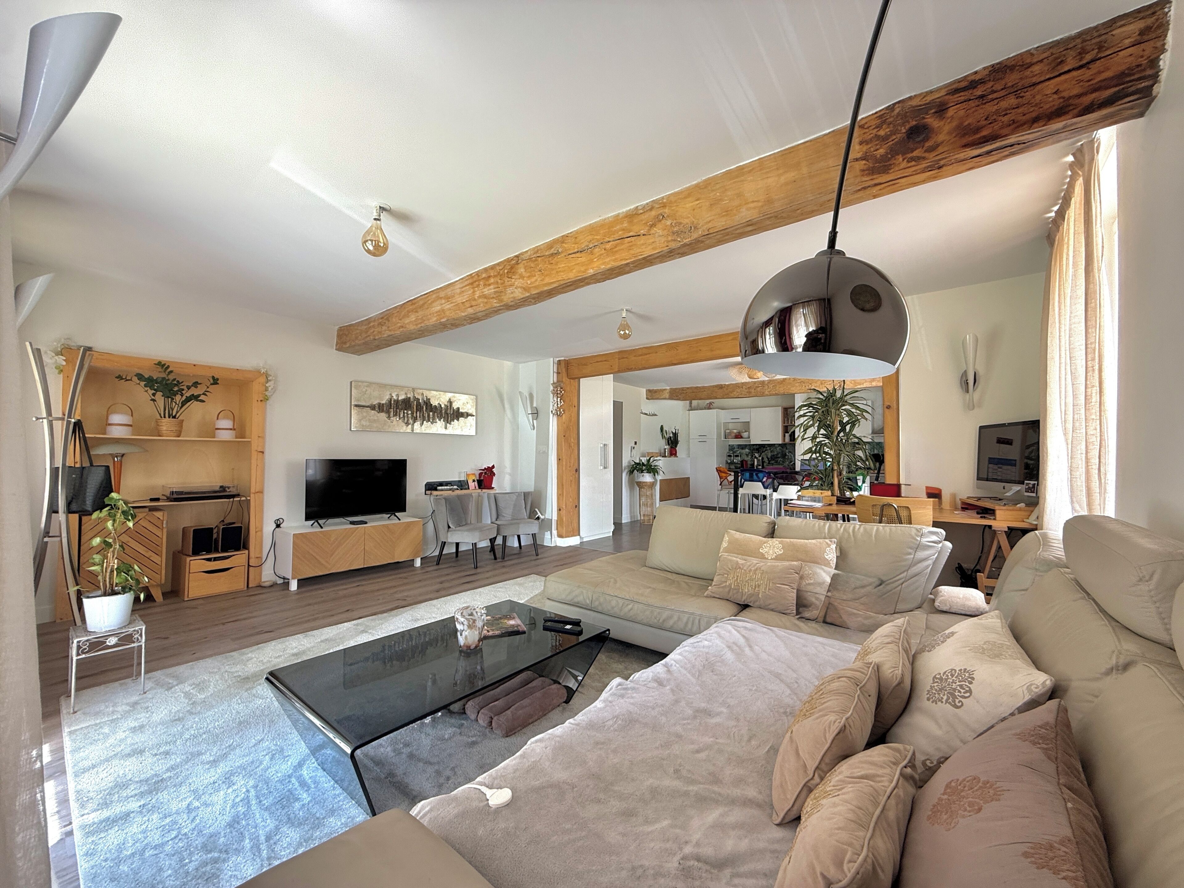 A modern living room with a sofa, coffee table, and TV. There is a wooden ceiling beam, a hanging light fixture, and a dining area in the background.