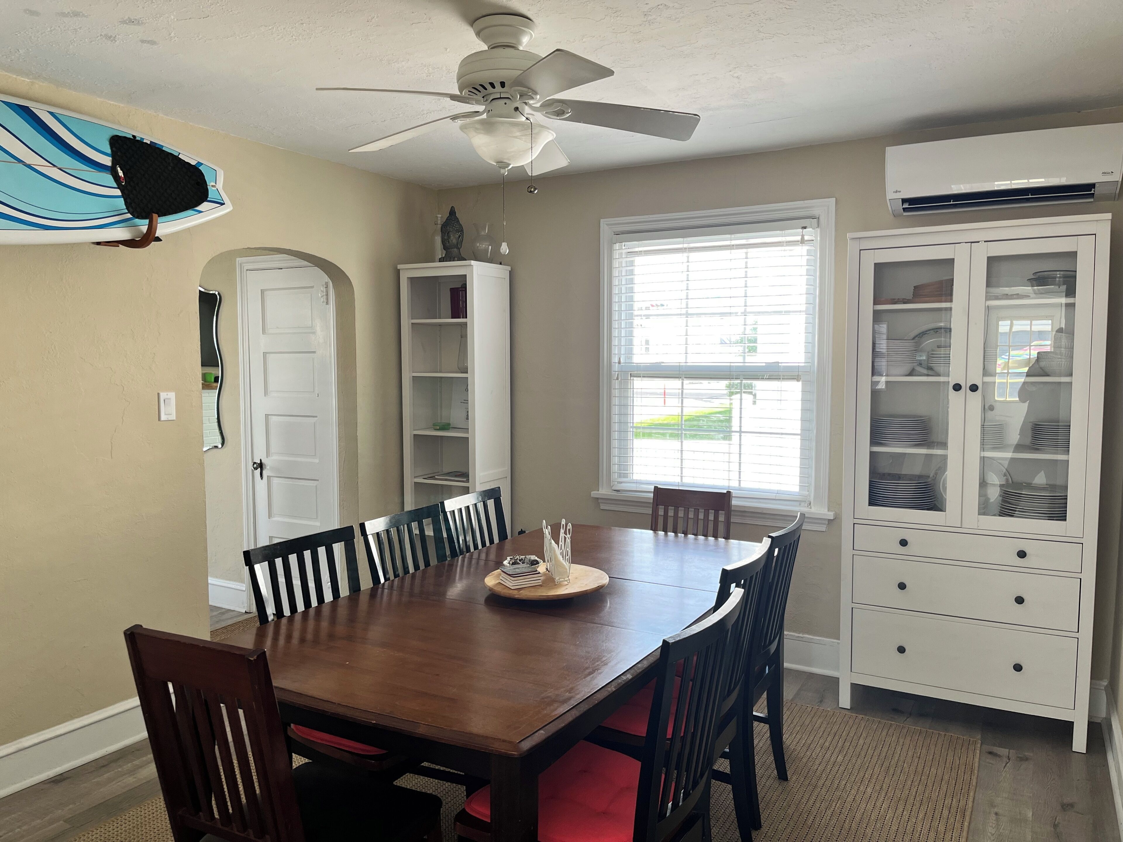 Une salle à manger avec une table en bois, des chaises, un ventilateur de plafond, une fenêtre avec des stores, un buffet blanc aux portes en verre et une planche de surf appuyée contre le mur.