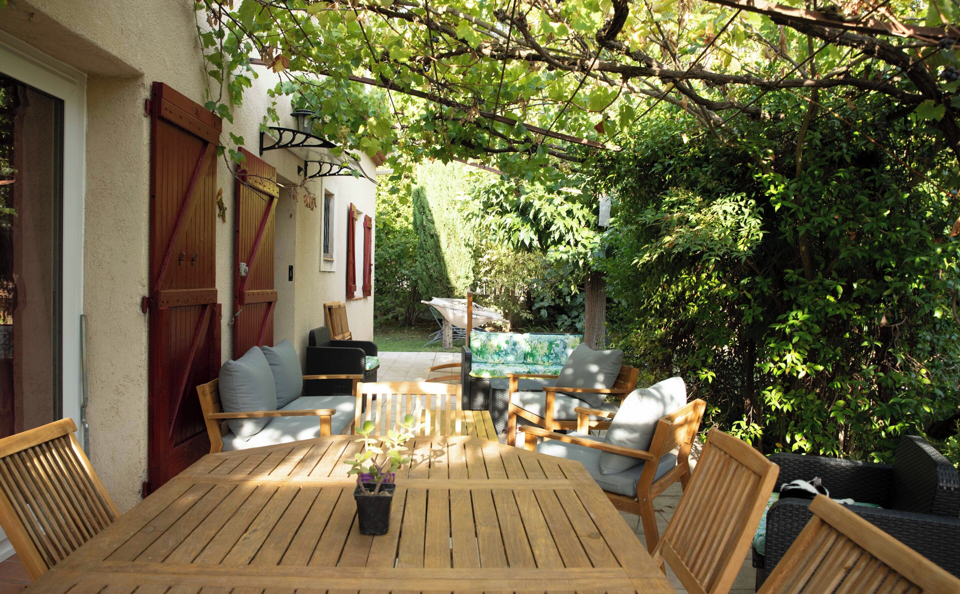 Un patio exterior con mesa y sillas de madera, rodeado de vegetación y vistas a un jardín.
