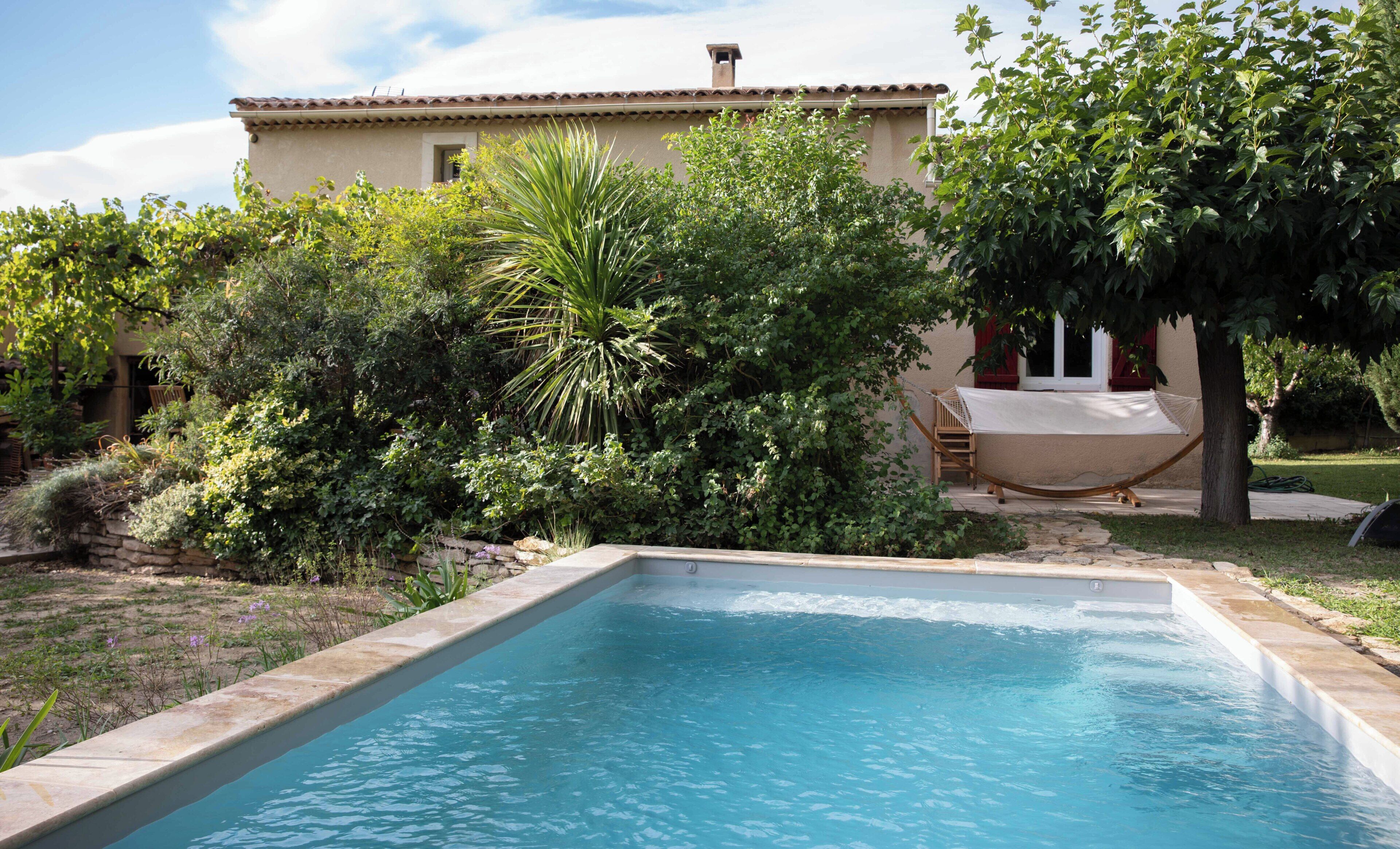 A swimming pool with a stone edge, surrounded by lush greenery and a building with a tiled roof in the background.