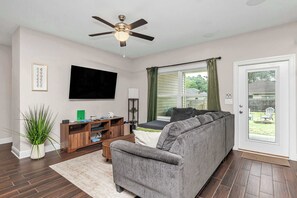 Ein Wohnzimmer mit grauem Sofa, einem Flachbildfernseher, einem Deckenventilator, einer Holzunterhaltungsecke und Blick ins Freie durch ein Fenster.