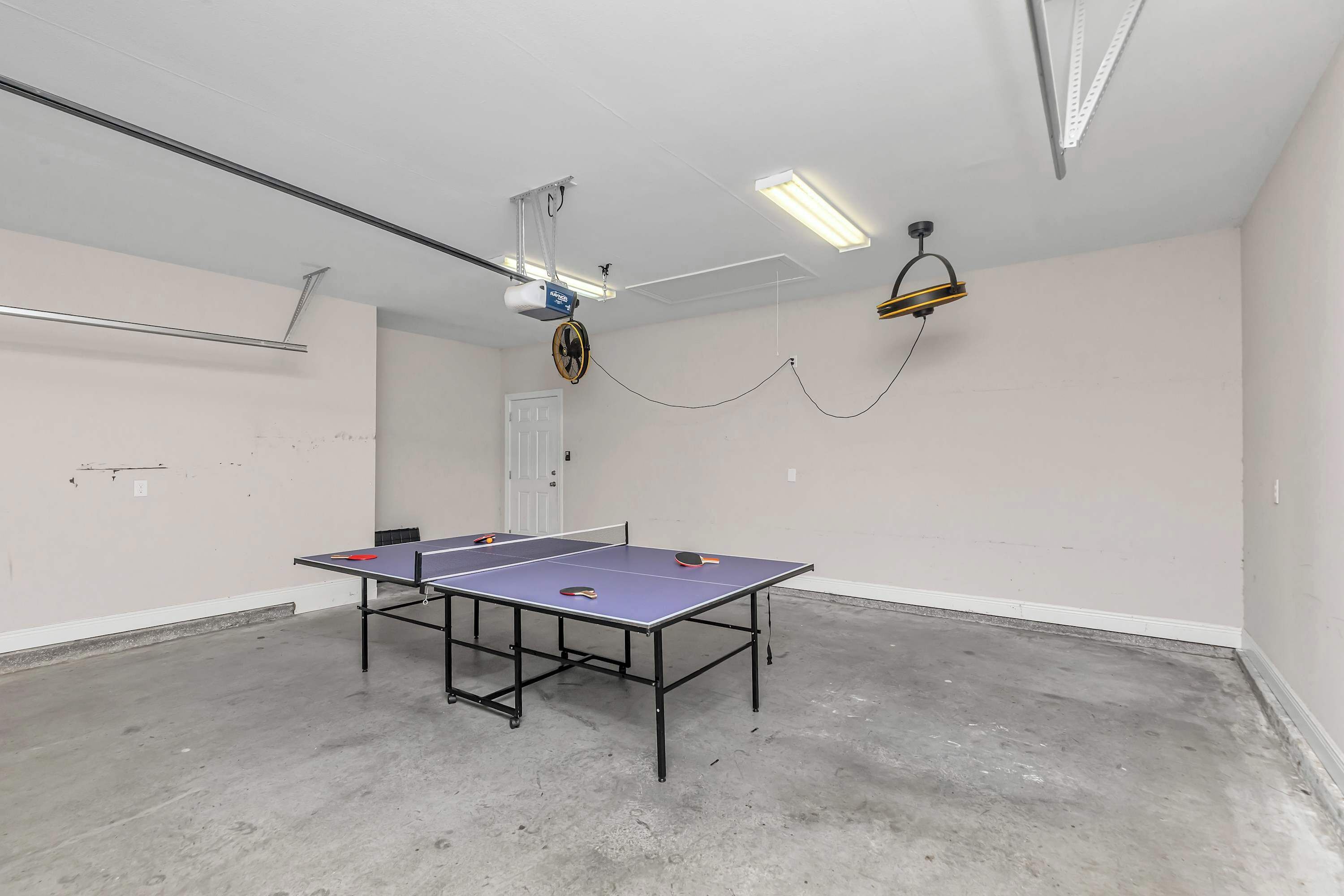 Una taula de ping-pong a una habitació amb ventilador de sostre, un projector i una làmpada penjant.