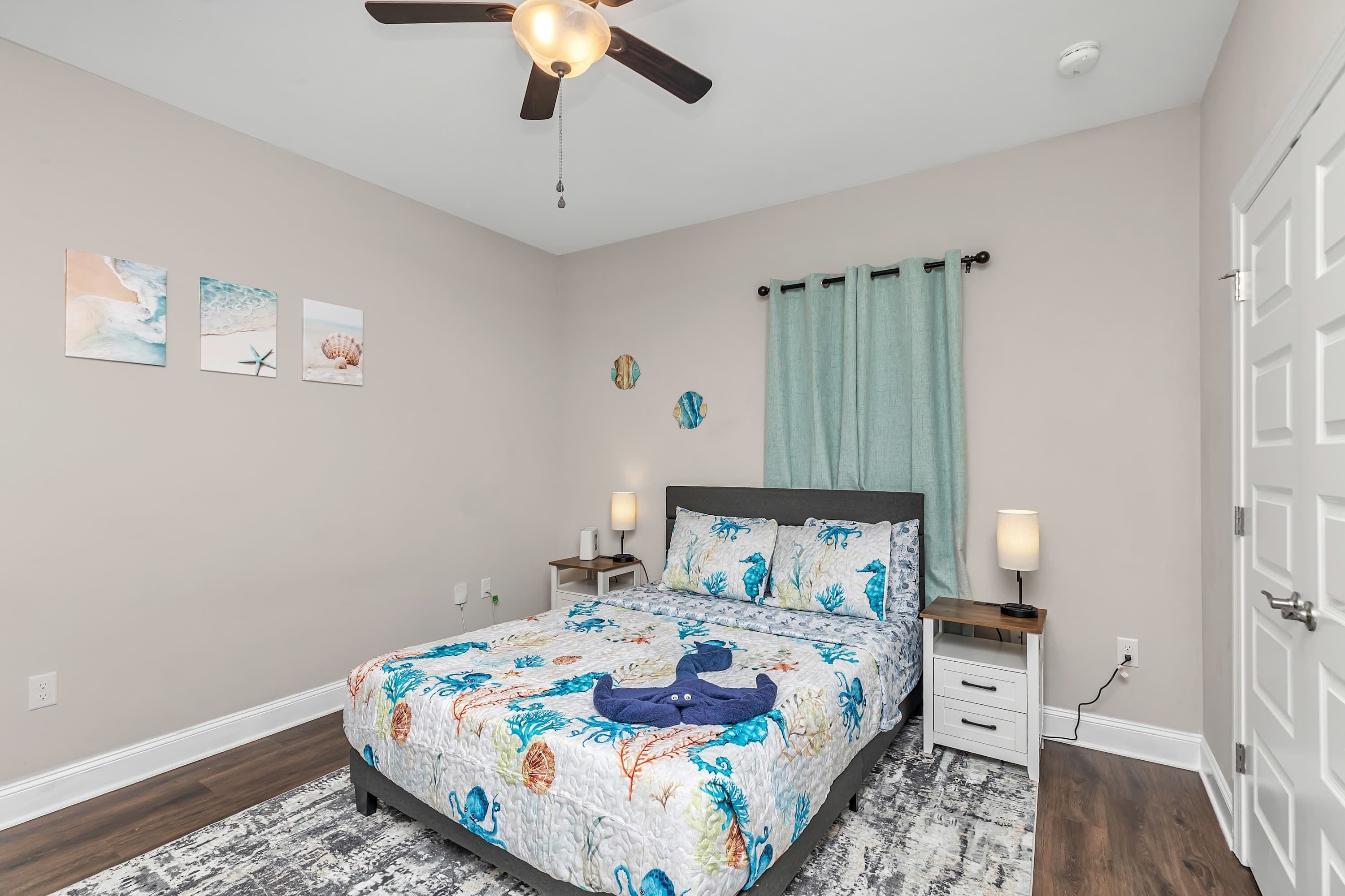 Un dormitorio con cama, mesitas de noche, ventilador de techo, ventana con cortinas y decoración en las paredes.