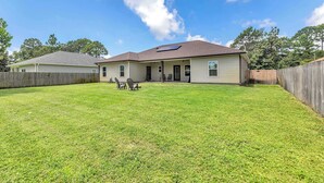 A single-story house with a fenced backyard, a patio with outdoor furniture, and a clear sky. - Oyster Cove: Pets Allowed, Grill, Pong, Beach! (Navarre)
