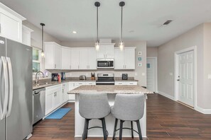 A modern kitchen with white cabinets, stainless steel appliances, and a central island with bar stools. - Oyster Cove: Pets Allowed, Grill, Pong, Beach! (Navarre)